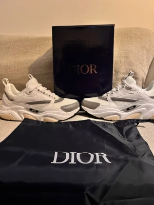 Vita Dior B22 sneakers - Snygga vita Dior B22 sneakers med gråa detaljer och chunky sula. Skorna har mesh och läderpaneler, samt tydlig Dior-logga på sidan. Perfekta för dig som gillar exklusiva och trendiga sneakers med modern design.