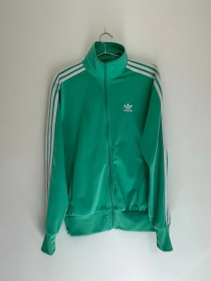 Grön Adidas track jacket - Snygg grön Adidas track jacket med klassiska vita ränder längs ärmarna och vit Adidas-logga på bröstet. Jackan har hög krage, dragkedja framtill och muddar vid ärmslut och midja. Tillverkad i glansigt syntetmaterial som ger en retro sportig look.