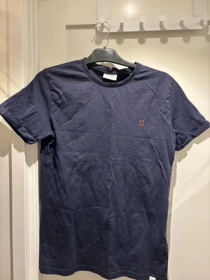 Mörkblå t-shirt från Les Deux | S - Snygg mörkblå t-shirt från Les Deux med liten röd broderad detalj på bröstet. Klassisk passform med rund hals och korta ärmar. Tillverkad i mjuk bomull som känns skön mot huden. Perfekt för en clean och stilren look.