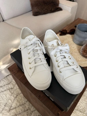 Vita Converse - Klassiska vita Converse sneakers i lågt utförande. Skorna har vita snören, guldiga öljetter och en vit gummisula med brunt mönster undertill. Tillverkade i canvas och passar perfekt till streetwear och casual outfits.