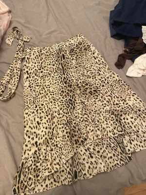 Leopardmönstrad omlottkjol med volang - Snygg leopardmönstrad omlottkjol med volangdetaljer och knytband i midjan. Kjolen har en asymmetrisk form och är perfekt för dig som vill sticka ut med ett trendigt djurmönster.