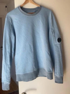 Ljusblå sweatshirt från C.P. Company - Ljusblå sweatshirt med rund hals från C.P. Company. Tröjan har en klassisk passform, ribbade muddar och en ikonisk svart linsdetalj på ena ärmen. Perfekt för en avslappnad och stilren look.