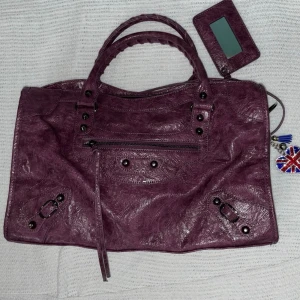 Lila Balenciaga handväska i skinn - Säljer en lila City handväska från Balenciaga i mjukt skinn med klassiska nitar och dragkedjedetaljer. Väskan har dubbla handtag, avtagbar spegel och en söt nyckelring med Union Jack. Perfekt för dig som gillar statement-accessoarer.