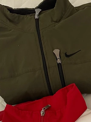 Olivgrön Nike vindjacka med dragkedja - Snygg olivgrön vindjacka från Nike med svart logga på bröstet och svarta dragkedjor. Jackan har hög krage och sportig design, perfekt för en streetwear-look. Materialet är lätt och vindtätt, vilket gör den grym för blåsiga dagar.