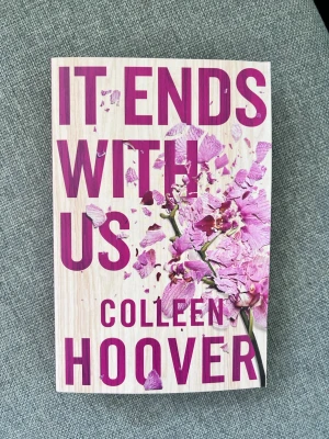 It Ends With Us av Colleen Hoover - En gripande roman av Colleen Hoover med omslag i träfärg och stora lila bokstäver samt en krossad lila blomma. Boken handlar om kärlek, svåra val och relationer. Perfekt för dig som gillar känslosamma berättelser och starka karaktärer.