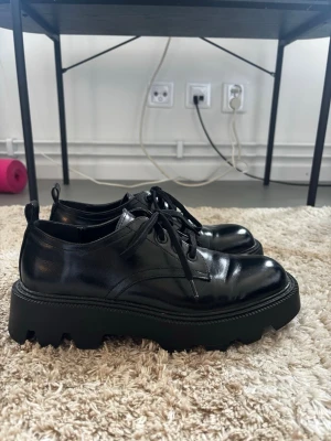 Chunky svarta loafers i skinn - Säljer ett par svarta chunky loafers med grov sula och snörning. Skorna är i blankt skinn och har en modern, edgy look som passar till många olika stilar. Fungerar toppen för mig som vanligtvis är en storlek 39!