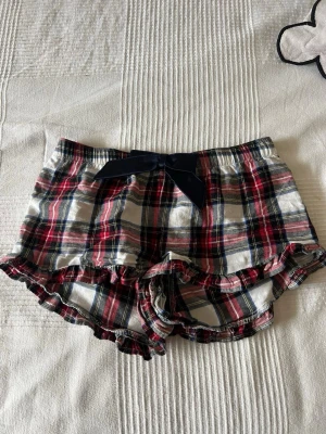 Rutiga pyjamasshorts med volang - Supersöta pyjamasshorts i röd, vit och marinblå rutig bomull med volangkant och en mörkblå rosett framtill. Perfekta för mysiga hemmakvällar eller sommarens varma nätter.