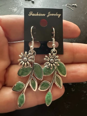 Silvriga örhängen med gröna blad - Unika örhängen i silverfärgad metall med hängande blommor och gröna blad. Blommorna har detaljerade kronblad och en markerad mitt, medan bladen har en glansig grön finish. Perfekta för dig som gillar naturinspirerade accessoarer.