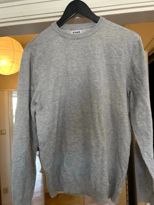 Ljusgrå stickad tröja  - En stilren ljusgrå stickad tröja med rund halsringning och långa ärmar. Tröjan har en klassisk passform och ribbstickade muddar vid ärmslut och nederkant. Perfekt att matcha med jeans, chinos eller kostymbyxor.
