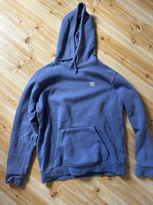 Lila hoodie från Adidas Originals - Lila hoodie från Adidas Originals med vit logga på bröstet. Klassisk modell med stor huva, dragsko och magficka. Mjuk bomullskänsla och ribbade muddar vid ärmslut och nederkant. Perfekt för en avslappnad streetstyle.