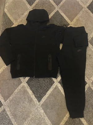 Svarta mjukisbyxor från Nike - Svarta mjukisbyxor från Nike med klassisk logga på benet. Byxorna har resår i midjan och muddar vid bensluten samt en praktisk ficka med lock på sidan. Materialet är mjukt och bekvämt, perfekt för en avslappnad stil.