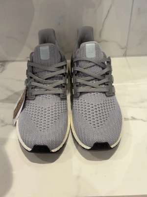 Grå Adidas Ultraboost sneakers - Storlek 45 sitter som ungefär 43,5. Snygga grå Adidas Ultraboost sneakers med stickad ovandel och klassiska tre ränder på sidan. Skorna har en vit Boost-sula för extra komfort och grå detaljer på häl och snörning. Perfekta för en sportig och trendig look.