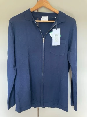 Marinblå merinoullskofta Lindbergh - Stilren marinblå stickad ziptröja från Lindbergh White i mjuk merinoull. Tröjan har hel dragkedja, klassisk krage och en bröstficka. Perfekt för lager-på-lager och enkel att matcha med andra plagg. Tröjan är helt oanvänd då jag har en liknande. Den är storlek L och sitter ganska tajt, skulle själv sätta storleken M/L lite beroende på hur man vill ha det.
