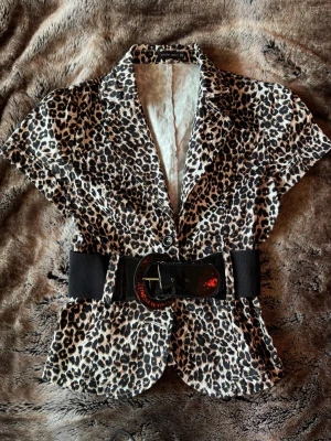 Leopardmönstrad blus med bälte - Snygg leopardmönstrad blus med korta ärmar och markerad midja tack vare ett brett svart bälte med stor spänne. Blusen har klassisk krage och knäppning framtill.  