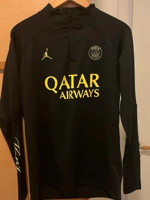 Svart PSG Jordan träningströja - Svart långärmad träningströja från PSG x Jordan med gul text 'Qatar Airways' på bröstet och Paris-logga på ärmen. Tröjan har halv dragkedja, PSG-emblem och Jordan-logga på bröstet. Tillverkad i ett lätt och sportigt material.