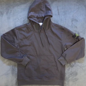Stone Island hoodie - Snygg mörkgrå hoodie från Stone Island med klassisk huva och dragsko. Har en stor magficka framtill och ribbade muddar vid ärmslut och nederkant. Ikonisk Stone Island-patch på vänster ärm. Perfekt för en avslappnad streetwear-look.