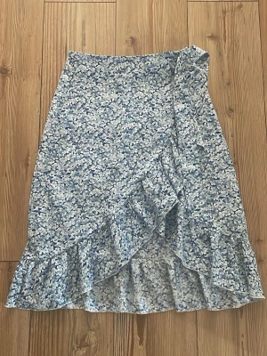 Blommig omlottkjol med volang - Söt blåvit omlottkjol med blommigt mönster och volangkant. Kjolen har en feminin och flowy look, perfekt för varma dagar. Lätt och luftig känsla med fina detaljer längs nederkanten.