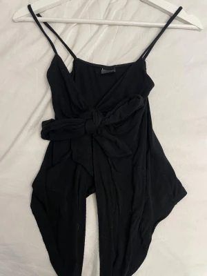 Svart jumpsuit med knytband - Svart linnetopp med smala axelband och brett knytband i midjan. Toppen är ärmlös, perfekt för en avslappnad och trendig stil. Tillverkad i mjukt bomullstyg som känns bekvämt mot huden.