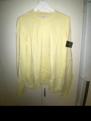 Gul långärmad tröja från Stone Island - Ljusgul långärmad topp från Stone Island med klassisk rund hals och ikonisk logopatch på ärmen. Tillverkad i mjuk bomull som känns skön mot huden. Perfekt för dig som vill ha en clean och stilren look med en touch av streetwear.