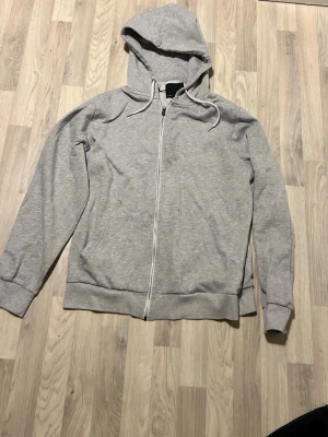 Ljusgrå zip-hoodie med huva - En stilren ljusgrå hoodie med dragkedja och huva. Tröjan har ribbade muddar vid ärmslut och nederkant samt två fickor framtill. Perfekt för en avslappnad och bekväm look. Dragsko i huvan för justerbar passform.
