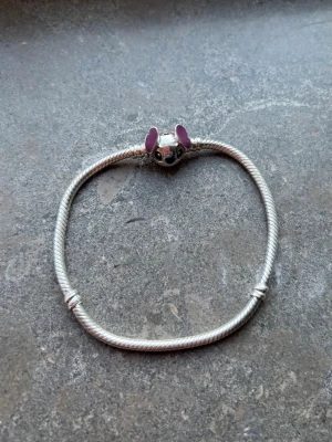 Pandora armband med Stitch-berlock - Säljer ett silverfärgat armband från Pandora med en söt Stitch-berlock. Berlocken har lila detaljer på öronen och armbandet har en klassisk tvinnad design. Perfekt för dig som gillar Disney och unika smycken.
