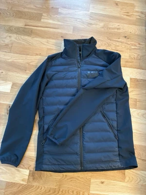 Svart pufferjacka från Helly Hansen - Snygg svart pufferjacka från Helly Hansen med quiltad front och rygg, höga krage och dragkedjor på fickorna. Jackan har elastiska muddar vid ärmsluten och diskret HH-logga på kragen. Perfekt för kalla dagar och enkel att matcha med allt.
