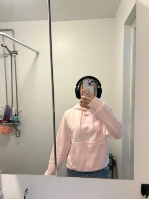 Ljusrosa hoodie från LYKO - Säljer en ljusrosa hoodie från LYKO med huva och magficka. Tryck med LYKO i rött framtill och stort på ryggen. Hoodien har dragsko i huvan och är gjord i mjukt material, perfekt för en chill och avslappnad stil.