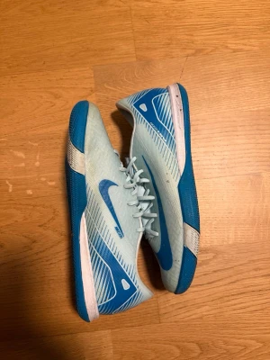 Nike blåa futsalskor - Knappt använda. Snygga ljusblåa Nike fotbollsskor för inomhusbruk med blå sula och vita detaljer. Skorna har snörning och en låg profil som ger bra grepp på inomhusplaner. Tillverkade i syntetmaterial med klassisk Nike-logga på sidan.