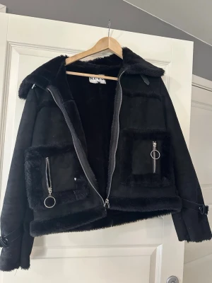 Svart pälsjacka från NA-KD - Snygg svart pälsjacka från NA-KD med stora fickor framtill, dragkedjor med ringdetaljer och bred krage. Jackan har fuskpälsdetaljer längs kanterna och på fickorna samt spännen vid ärmsluten. 