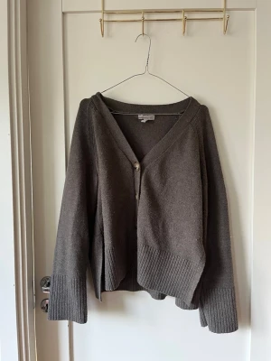 Brun stickad kofta - Mysig brun kofta med v-ringning och en dekorativ knapp framtill. Koftan har långa ärmar och bred ribbstickad kant vid ärmslut och nederkant. Perfekt för lager på lager och enkel att matcha med olika outfits.