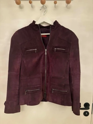 Vinröd jacka med dragkedjor - Snygg vinröd jacka med fyra dragkedjefickor framtill och ståkrage. Jackan har en rak passform och stängs med dragkedja. Perfekt för dig som vill ha en cool och stilren look.