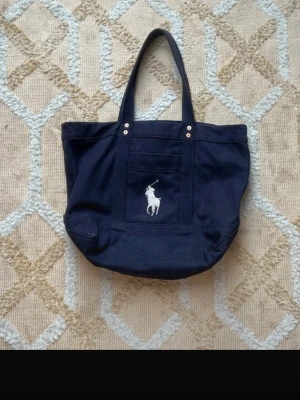 Mörkblå shoppingväska från Polo Ralph Lauren - Snygg mörkblå shoppingväska i canvas från Polo Ralph Lauren med klassisk vit broderad logga framtill. Väskan har dubbla handtag och en praktisk ytterficka. Perfekt för att bära med sig allt man behöver under dagen.