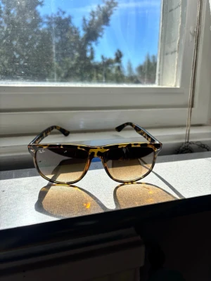 Ray-Ban solglasögon leopardmönster - Snygga Ray-Ban solglasögon med klassisk fyrkantig form och leopardmönstrad båge i bruna och gula toner. Glasen är tonade och ger en cool retrovibe. Perfekta för att lyfta din stil under soliga dagar.