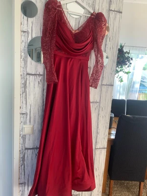 Vinröd långklänning med spetsärmar - Elegant vinröd långklänning med långa spetsärmar och draperad urringning. Klänningen har en figurnära överdel och ett flytande kjolparti i glansigt material. Perfekt för bal eller festliga tillfällen.