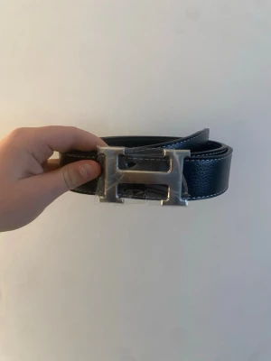 Svart läderbälte med H-spänne - Snyggt svart bälte i läder med ett stort, silverfärgat H-format spänne. Bältet har vita kontrastsömmar längs kanterna och flera hål för justerbar passform. Perfekt accessoar för att lyfta vilken outfit som helst.