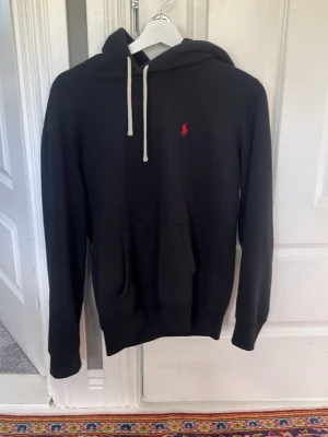Svart Ralph lauren hoodie - Svart hoodie från Ralph Lauren med klassisk röd logga broderad på bröstet. Hoodien har huva med vita snören och en stor magficka framtill. Det är bara att hör av er om ni undrar något.