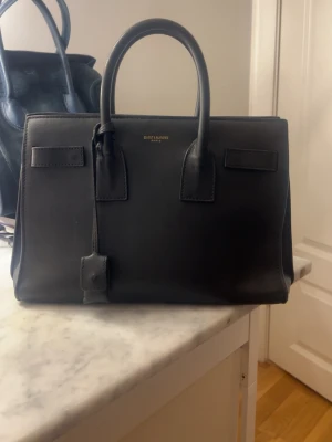 Svart handväska från Saint Laurent - Snygg svart handväska från Saint Laurent i slätt läder med två topphandtag och guldfärgade dragkedjor. Klassisk boxig form och diskret logga framtill. Perfekt för dig som gillar stilrena och lyxiga accessoarer.