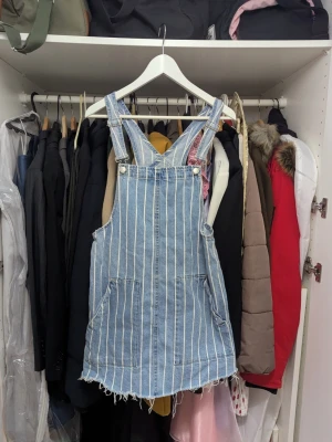 Randig jeans klänning med hängslen - Snygg jeans klänning i ljusblå denim med vita vertikala ränder. Modellen har klassiska hängslen, två stora fickor framtill och rå, fransad nederkant för en avslappnad look. Perfekt för en trendig och avslappnad stil.