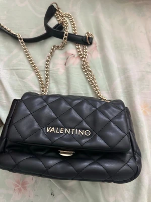 Svart quiltad axelväska Valentino - Snygg svart axelväska från Valentino med quiltat mönster och guldfärgad kedjerem. Väskan har ett lock med magnetlås och logotyp i guld framtill. Tillverkad i mjukt konstläder och har flera fack inuti för enkel organisering.