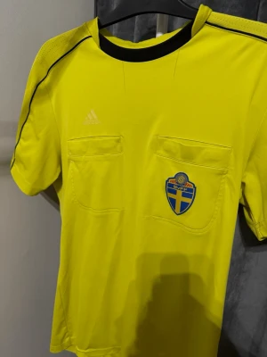 Gul domartröja SvFF Adidas - Gul domartröja från Adidas med SvFF-emblem på bröstet och Fair Play/Svenska Spel-tryck på ärmen. Tröjan har två bröstfickor, svarta detaljer vid axlar och krage samt är tillverkad i funktionsmaterial. Perfekt för fotbollsdomare.