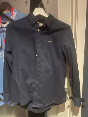 Mörkblå skjorta från Gant - Stilren mörkblå skjorta från Gant med klassisk krage och vita knappar. Skjortan har lång ärm och en liten broderad logga på bröstet. Perfekt för dig som gillar en clean och tidlös look.