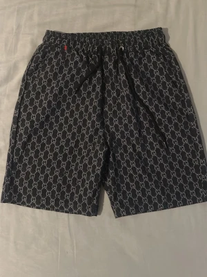 Svarta shorts med mönster - Storlek XS men passar även SSnygga svarta shorts med klassiskt GG-monogram över hela tyget. De har elastisk midja med dragsko och sidofickor. Perfekta för en trendig och avslappnad stil. Materialet känns lätt och bekvämt.