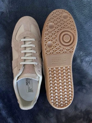 Mason margiela gats 42 - Stilrena sneakers från Maison Margiela i beige mocka och läder med vita skosnören. Klassisk låg modell med gummisula och diskret branding på sulan. Perfekta för dig som gillar minimalistisk och trendig design.
