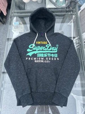 Superdry hoodie – mörkgrå vintage print (S) - Säljer en riktigt clean Superdry hoodie i mörkgrå melerad färg med klassiskt vintage print 🔥  Perfekt för sommarkvällar, sena utekvällar eller bara en clean streetwear fit. Sjukt enkel att styla – passar till både jeans, shorts och cargos.  • Storlek: S • Skick: mycket bra (inga hål eller fläckar) • Material: bomull/polyester-blandning • Fleece på insidan = mjuk & bekväm • Normal passform  Sitter riktigt nice och funkar året runt – en sån basic som alltid går hem.