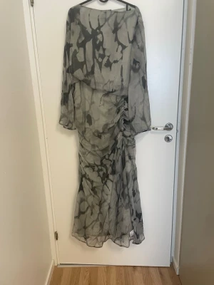 Klänning  - Säljer en lång, transparent maxiklänning i grått med svart abstrakt mönster. Klänningen har långa, vida ärmar och rynkade detaljer längs sidan för en cool och trendig look. Perfekt för dig som gillar unika plagg med flowy känsla.