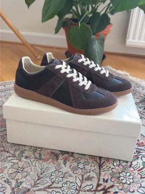 Maison Margiela Replica sneakers brun - Snygga Maison Margiela Replica sneakers mörkbrunt och svart läder med mockadetaljer. Skorna har vita snören och en klassisk gummisula i ljusbrunt. Perfekt för dig som gillar stilrena och tidlösa sneakers med premiumkänsla. Storlek 43 