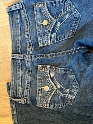 Blå bootcut jeans med broderade fickor - Snygga blå bootcut jeans med broderade detaljer och paljetter på bakfickorna. Jeansen har klassisk femficksdesign, gylf med dragkedja och knapp samt bälteshällor. Perfekta för dig som gillar en lite utsvängd modell och vill sticka ut med detaljer. Kontakta för att diskutera pris. Nypris 350kr tror jag 