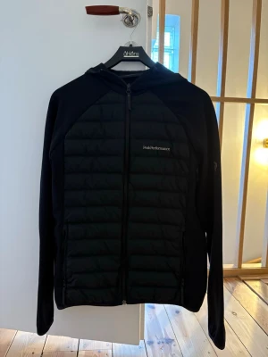Peak Performance cardigan  - Svart pufferjacka från Peak Performance med quiltad front, hel dragkedja och huva. Jackan har diskret logga på bröstet och en liten logga på ärmen som har litet slitage men annars i jättefint skick! Storlek L men passar som M.