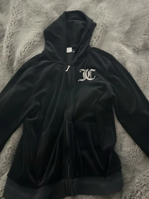 Svart velourhoodie Juicy Couture - Svart hoodie i mjuk velour från Juicy Couture med dragkedja framtill. Stora glittriga bokstäver 'JC' på bröstet och 'Juicy Couture' på ryggen. Klassisk huva och långa ärmar, perfekt för en trendig och avslappnad look.