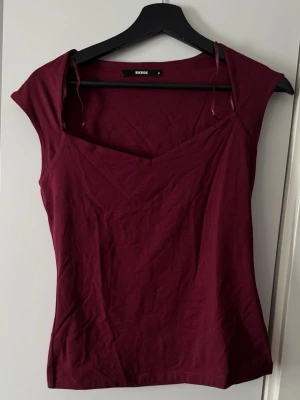 Vinröd topp med v-ringning från BikBok - Vinröd topp från BikBok med v-ringad hals och smala axelband. Modellen är ärmlös och har en figurnära passform. Tillverkad i mjuk bomull som känns skön mot huden. Perfekt för att skapa en stilren och trendig look.